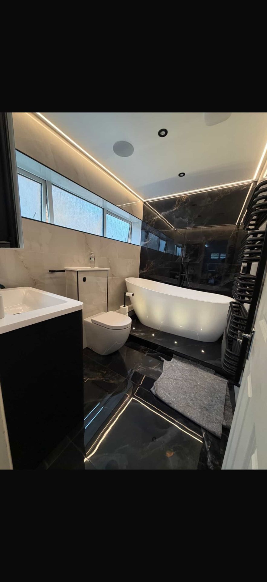 Dark marble bathroom with LED ceiling strip lighting, freestanding bath and floor-level plinth lights