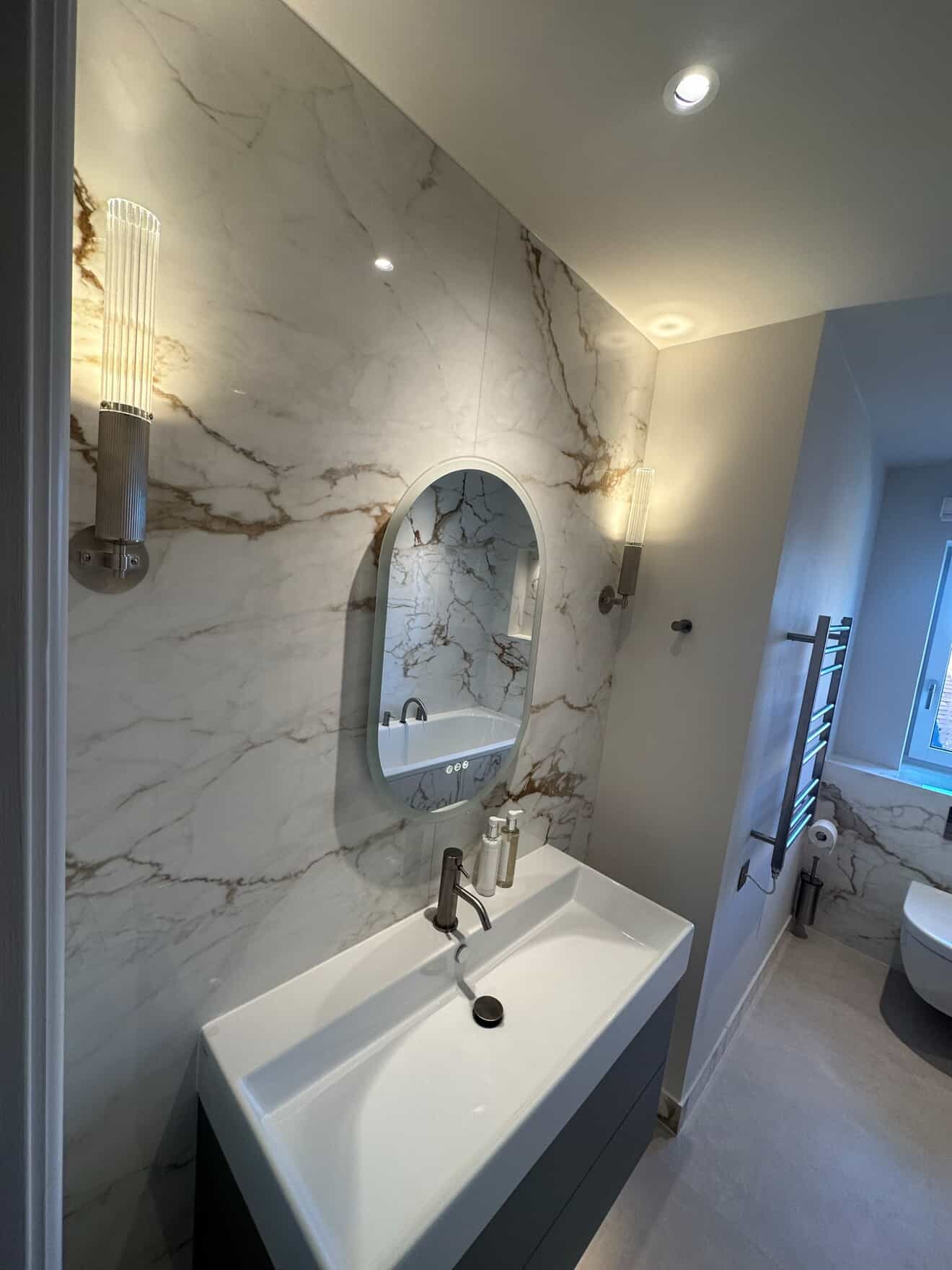 Bathroom with marble feature wall, oval backlit mirror and decorative wall sconces